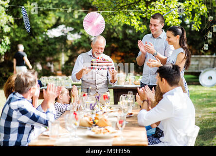 Fête de famille à l'extérieur dans la cour. Big garden party. Fête d'anniversaire. A senior man holding a gâteau d'anniversaire. Banque D'Images