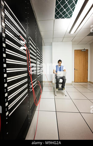 Technicien en informatique travaillant sur un portable tout en faisant le travail de diagnostic sur un serveur dans une plus grande salle de serveurs informatiques. Banque D'Images