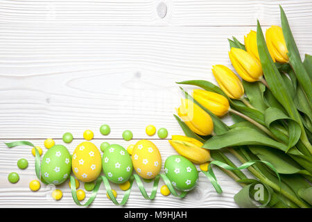 Oeufs de Pâques en chocolat, bonbons et tulipes jaunes sur fond de bois blanc. Banque D'Images