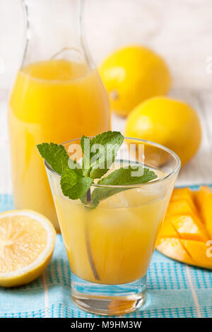 Limonade à la mangue avec une branche de menthe dans un verre sur une serviette bleue, mangue fraîche et citrons sur un fond de bois blanc Banque D'Images