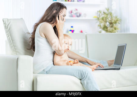 Jeune mère tenant un bébé de l'année dans ses bras,parler sur son téléphone cellulaire et working on laptop Banque D'Images
