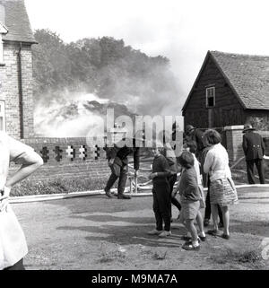 Années 1960, historiques, des pompiers avec flexibles assister à un incendie dans la cour de certains bâtiments agricoles,avec les enfants et les personnes se trouvant sur l'observation, Buckinghamshire, Angleterre, Royaume-Uni. Banque D'Images