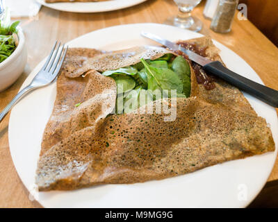 Salé crepe sarrasin rempli d'spinich sur une plaque blanche : une délicieuse crêpe mince brun est soigneusement replié autour d'un lit d'obturations surmontée de bébé vert spinish sur une plaque blanche avec couteau et fourchette. Banque D'Images