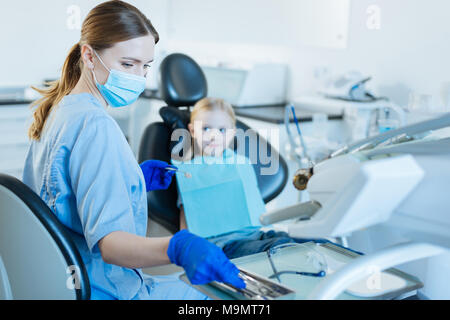 Bilan rapide. Charmante jeune femme dentiste tenant un miroir de la bouche et en tenant une sonde bouche nécessaire à l'examen de son petit patient Banque D'Images