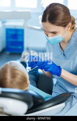 Médecin doux. Agréable Jeune femme tenant une sonde dentaire dentiste et un miroir de la bouche et d'être sur le point d'effectuer un bilan de ses petits patients o Banque D'Images