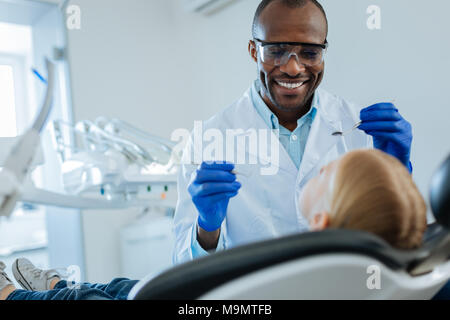 Sourire encourageant. Cheerful male dentist tenant un miroir de la bouche et une sonde dentaire et souriant à son petit patient pendant l'exécution de cavité buccale e Banque D'Images