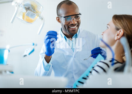 Conversation agréable. Beau male dentist holding instruments spéciaux et de discuter avec son patient de sexe féminin avant d'effectuer un examen checkup o Banque D'Images