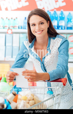 Smiling woman doing courses au supermarché, elle s'appuie sur un panier et la tenue d'une liste Banque D'Images