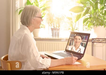 Hauts femme dans la salle de séjour ensoleillée en face d'un ordinateur portable prendre des notes pendant un appel vidéo avec sa femme médecin Banque D'Images