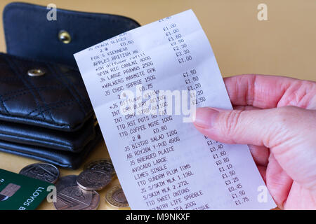 Femme main tenant une épicerie supermarché ticket de caisse avec un sac à main, quelques pièces de monnaie et carte bancaire dans l'arrière-plan, Royaume-Uni Banque D'Images