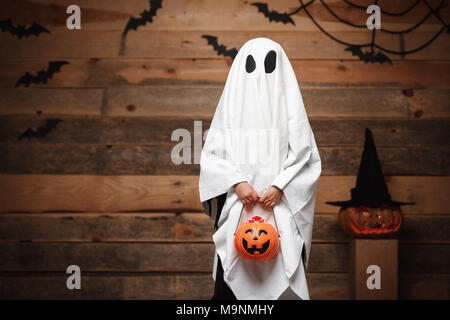 Concept d'Halloween - peu de blanc fantôme avec citrouille halloween candy jar faisant le tour ou les traiter avec des citrouilles et des chauves-souris sur spider web studio en bois sur l'arrière-plan. Banque D'Images