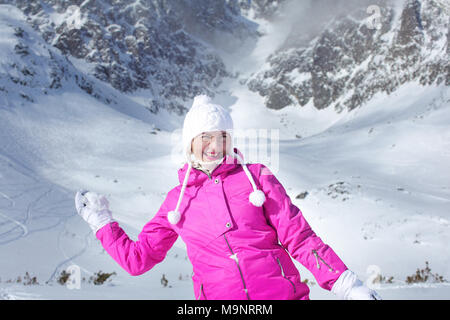 Happy smiling young woman in pink veste de ski se préparent à lancer une boule de neige avec la montagne enneigée en arrière-plan. Banque D'Images