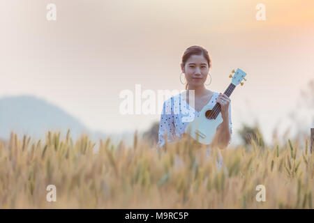 Belle asian woman holding ukulele et détente à champ d'orge en été l'heure du coucher de soleil. Banque D'Images