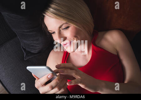 Une femme heureuse dans un T-shirt et pantalon gris se trouve à la maison sur le canapé dans la soirée et est titulaire d'un téléphone mobile. Une femme est shopping par téléphone. En ligne Banque D'Images