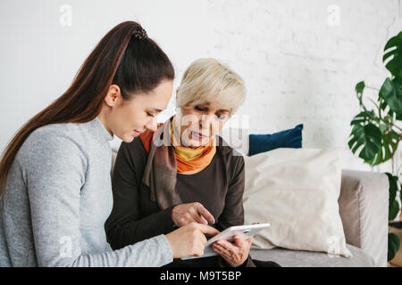 Une jeune fille bénévole ou petite-fille explique à une femme âgée comment utiliser une tablette ou manifeste une certaine application ou vous enseigne comment utiliser un Banque D'Images