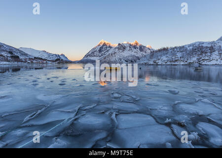 Scène d'hiver de bateau dans le fjord partiellement gelé et paysage enneigé. La glace et l'eau bleue avec des reflets de sommets de montagnes en orange au coucher du soleil. Banque D'Images