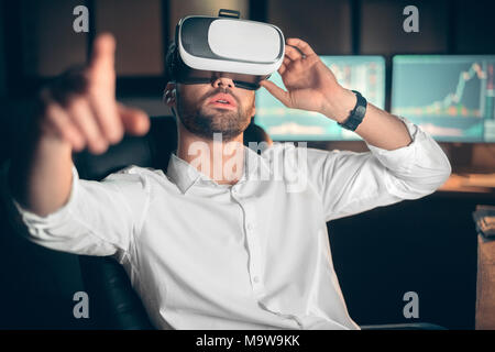 Jeune homme barbu trader au bureau assis à table le port casque de réalité virtuelle pointant concentré de l'appareil photo Banque D'Images