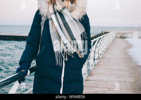 Jeune femme se dresse sur la promenade d'hiver contre la mer, vêtu d'une veste vers le bas, hotte, gants et écharpe. Marcher dans la saison froide dans des vêtements chauds Banque D'Images