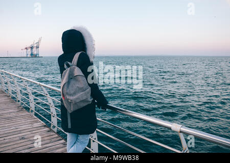 Jeune femme dans le capot à l'hiver à la mer. Vue arrière de meilleur dans des Down jacket avec un sac à dos debout sur le bord de l'eau ressemble au w Banque D'Images