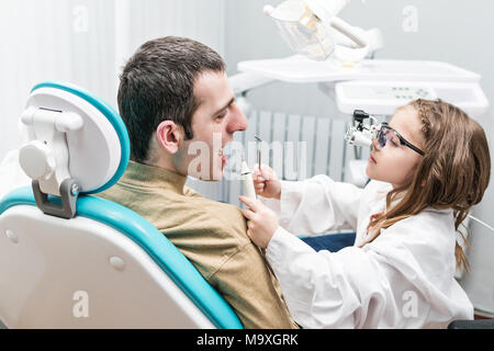 Petite fille dans une robe blanche dans l'image d'un dentiste examine les dents pour un homme assis dans un fauteuil dentaire Banque D'Images