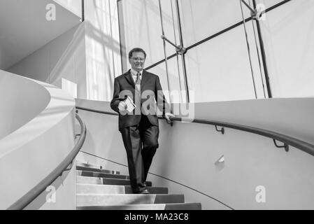 Vieil homme vêtu d'un costume à un escalier vers le bas d'un immeuble avec grande fenêtre en verre ; noir et blanc Banque D'Images