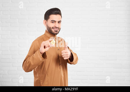 Les jeunes musulmans attrayant modèle masculin dans les vêtements islamiques d'être heureux de présenter et bitcoin or Thumbs up in front of blurred white brick wall. Plan horizontal. Banque D'Images