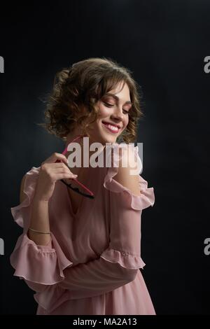 Façade de jolie jeune fille brune aux cheveux bouclés, qui n'est sourire, rire et garder les lunettes à la mode. Modèle porte robe rose avec ouvert aux épaules et lumière jour composent. Banque D'Images