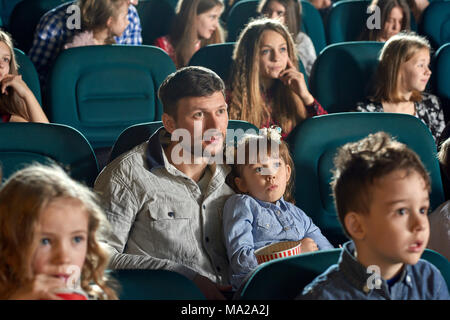 Photo de père avec petite fille regarder la vidéo dans le cinéma. Jeune fille est à la recherche très mignons et intéressés, eating popcorn. Il y a d'autres personnes est sorti sur l'arrière-plan. Banque D'Images