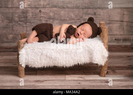 Deux semaines de bébé nouveau-né garçon portant un ours brun, moufles, bonnet. Il est en train de dormir sur un petit lit en bois, et de câliner un animal en peluche. Banque D'Images