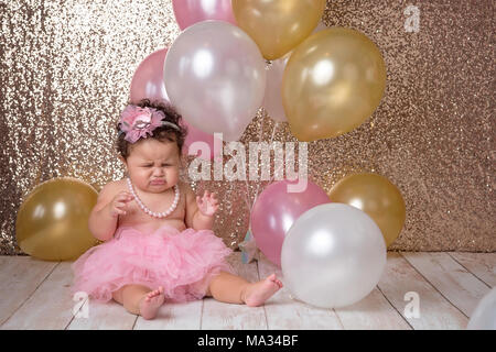 Un malheureux, un an, bébé fille assise avec un bouquet de ballons. Il est vêtu d'un tutu rose et un collier de perles. Banque D'Images