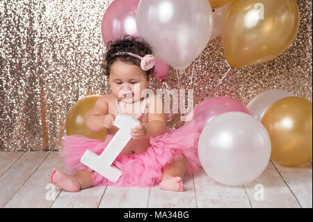 A un an, bébé fille jouant avec un grand bloc de la forme d'un numéro un. Il est vêtu d'un tutu rose, fleur bandeau et un collier de perles. Banque D'Images