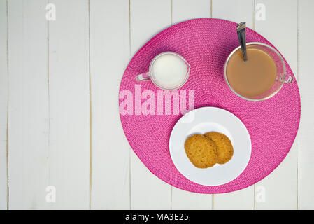 Du thé chaud, des biscuits et du lait sont disposés sur un napperon rose sur un tableau blanc, tableau. Le plateau a lait en elle. L'arrangement est décentré et tourné à partir de Banque D'Images