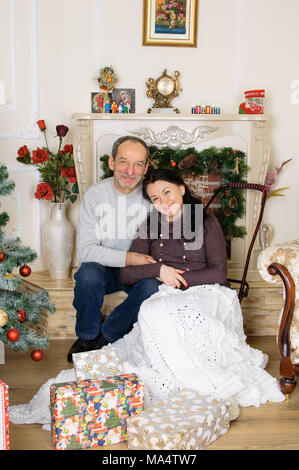 Studio Portrait de couple avec différence d'âge assis près de cheminée artificielle et l'arbre de Noël avec les coffrets cadeaux et boules rouges et Hugging Banque D'Images
