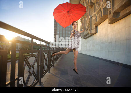 Ivano-Frankivsk, Ukraine - 1 juin 2015 : young pretty dancing girl saute, holding heart-shaped parapluie rouge. Il est intéressant de façade du bâtiment ville texturé sur arrière-plan. Banque D'Images