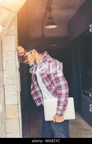 Hipster déprimé avec l'homme serra les poing posant sa tête contre un mur avec ordinateur portable à la main. Banque D'Images