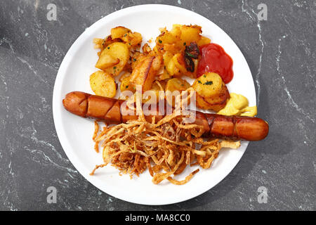 Close up partie d'une grande saucisse grillée avec des pommes de terre rôties, oignons frits, ketchup et moutarde sur plaque blanche sur le tableau gris, Eleva Banque D'Images