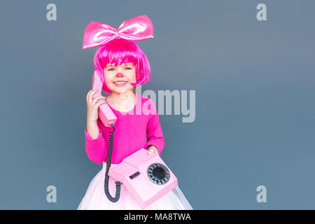 Portrait de petite fille clown appelant les vieux téléphone rose, souriant à pleines dents. Studio shot, isolé sur fond gris Banque D'Images