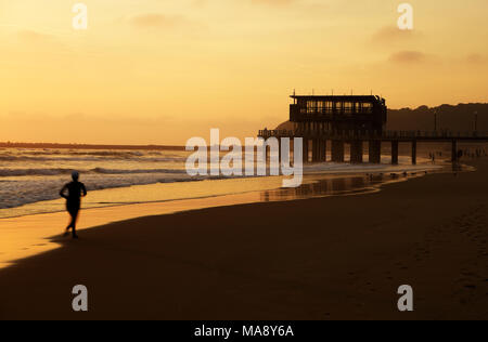 Silhouette de jeune femme en marche le long du rivage de l'uShaka plage sur le front de mer du Golden Mile de Durban, Afrique du Sud, au lever du soleil Banque D'Images