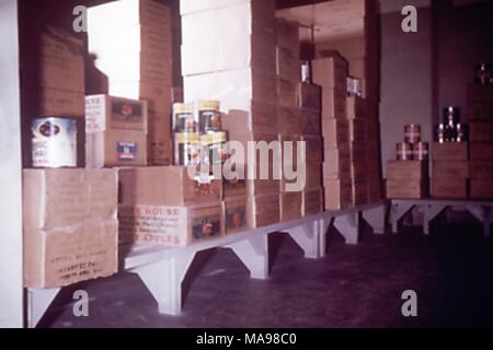 Photo d'une zone de stockage alimentaire intérieure à un camp de travail des travailleurs migrants, avec étagères bordées de boîtes en carton brun et de grandes boîtes de conserves de pommes, 1975. Image courtoisie CDC. () Banque D'Images