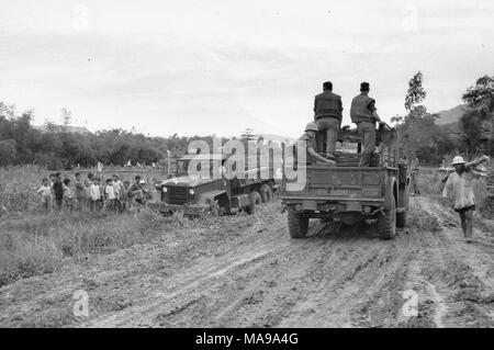 Photographie noir et blanc montrant un groupe de trois soldats américains, école loin dans le lit ouvert d'une camionnette, deux vers l'avant, l'un, situé sur une route de terre dans la campagne, avec un autre camion et les gens à gauche, et à droite en bambou personne exerçant, photographié au Vietnam pendant la guerre du Vietnam, 1971. () Banque D'Images