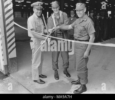 Photographie en noir et blanc de trois ans, des militaires américains, l'un un Navy Seabee représentant, Comité permanent, en vue de pleine longueur, tenant une paire de ciseaux géants, prêt à couper un ruban tendu à travers l'entrée d'un entrepôt d'approvisionnement, avec d'autres soldats en regardant en arrière-plan, photographié au Vietnam pendant la guerre du Vietnam (1955-1975), 1968. () Banque D'Images