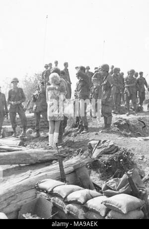 Photographie noir et blanc montrant un grand groupe de soldats américains en uniforme, un soldat avec son bras autour d'une femme blonde qui porte une mini-robe à motifs, des bottes et la tenue d'un sac à main, tous debout dans un paysage rocheux, avec trous de sacs et d'armes à l'avant-plan, photographié au Vietnam pendant la guerre du Vietnam (1955-1975), 1971. () Banque D'Images