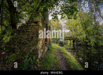 Un petit chemin ombragé, calme, passe à côté d'un canal et les vestiges de pierre d'une ancienne ferme, et mène à une petite passerelle en bois au loin. Banque D'Images