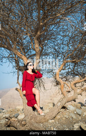 Belle jeune fille en robe rouge sous un arbre dans le désert Banque D'Images