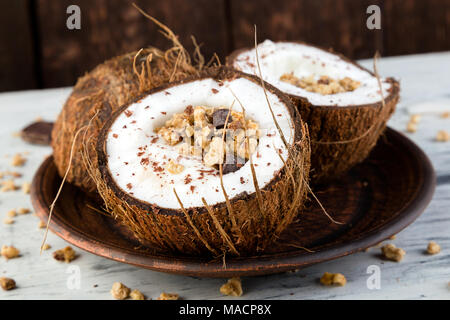 Petit-déjeuner sain dans la noix de coco noeud sur fond blanc. Le yogourt dans un bol de noix de coco avec des flocons de noix de coco, chocolat et muesli. Vue de dessus, des laïcs, des frais généraux Banque D'Images