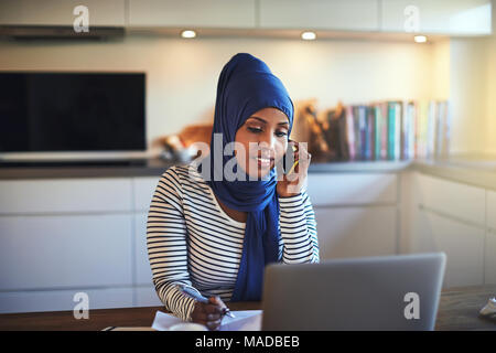 Jeune femme entrepreneur arabe portant un hijab assis à sa table de cuisine l'utilisation d'un cellulaire et à l'aide d'un ordinateur portable tout en travaillant de chez vous. Banque D'Images