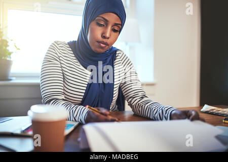L'accent jeune musulman femme entrepreneur portant un hijab lire la paperasse tout en travaillant à table dans son bureau à domicile Banque D'Images