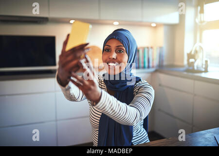 Souriante jeune femme arabe portant un hijab assis dans sa cuisine à la maison pour s'autoportraits avec son portable Banque D'Images