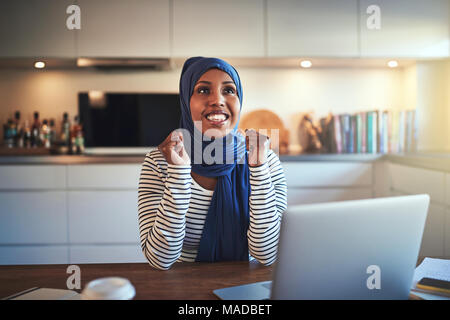 Jeune femme entrepreneur musulman extatique portant un hijab Célébrons les succès tout en étant assis à une table dans sa cuisine et de travailler sur un ordinateur portable Banque D'Images