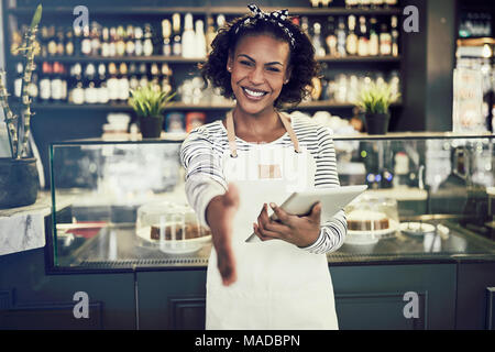 Smiling young African cafe owner s'étendant d'une poignée de main tout en se tenant dans un café branché tenant une tablette numérique Banque D'Images